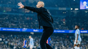 A bald man in a black jacket gestures passionately during a soccer match with players from Manchester City visible in the background playing in UEFA Champions League.