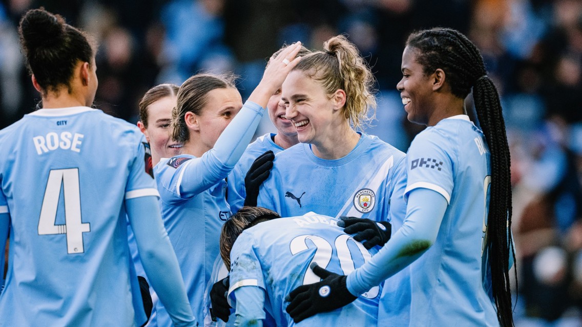 A Manchester City Women team huddle on the field, wearing their light blue kits with prominent logos. Players gather in a circle showing team solidarity.