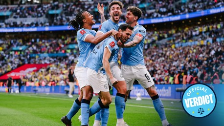 Manchester City players celebrating a goal during a football match at Wembley Stadium with a crowd in the background and an 'Official Verdict' badge visible.