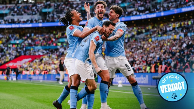 Manchester City players celebrating a goal during a football match at Wembley Stadium with a crowd in the background and an 'Official Verdict' badge visible.