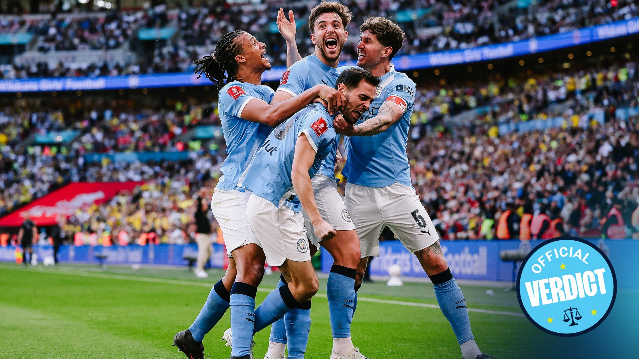 Manchester City players celebrating a goal during a football match at Wembley Stadium with a crowd in the background and an 'Official Verdict' badge visible.