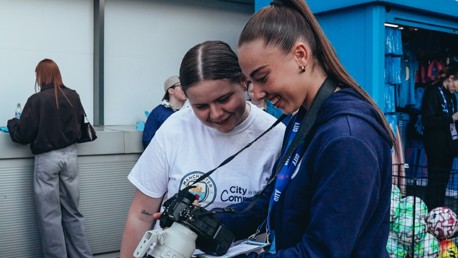 Two women reviewing photos on a camera outside a blue booth with merchandise in the background.