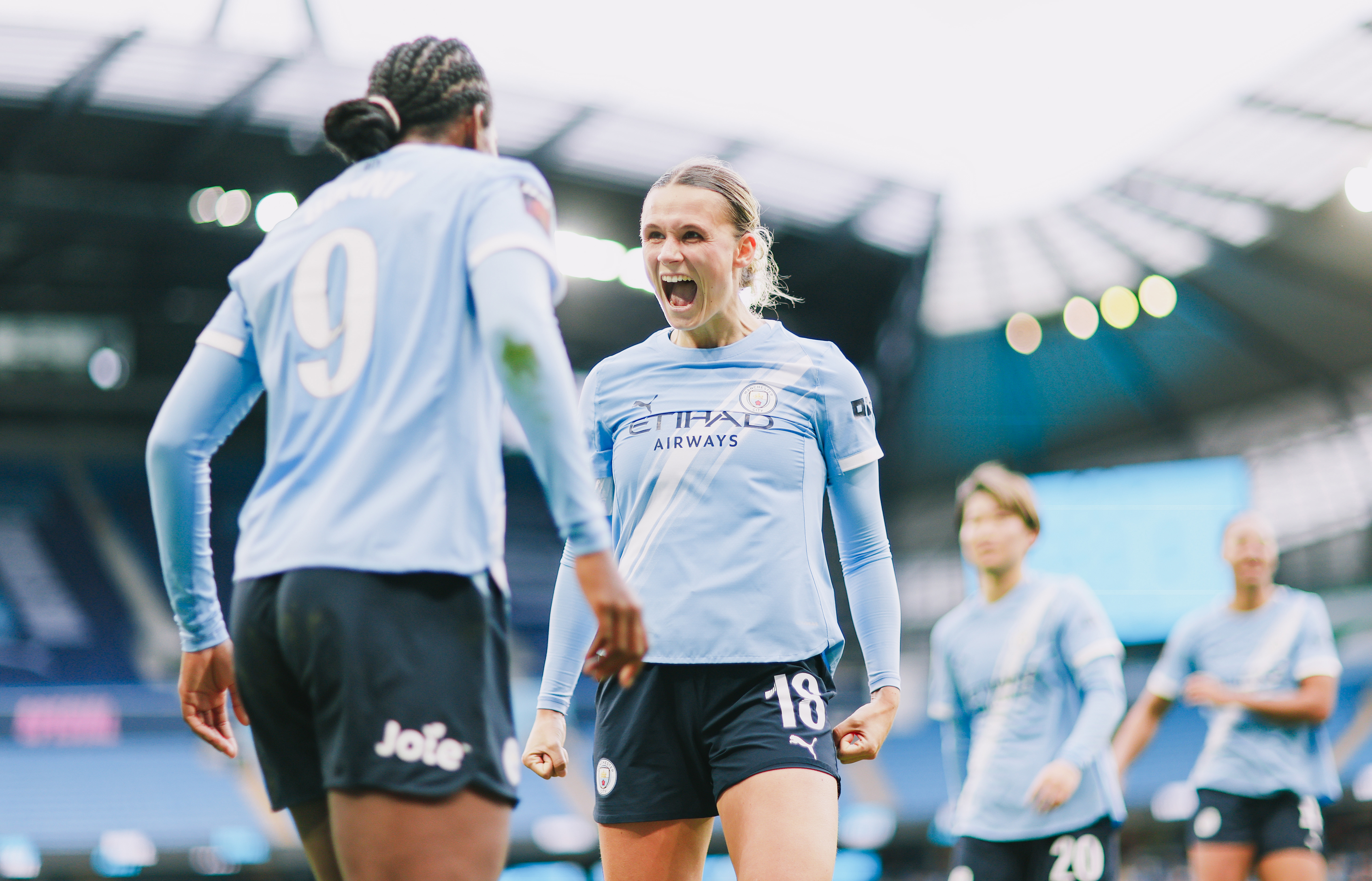 Women's football players celebrating on the field in Manchester City kits after scoring a goal at the Etihad Stadium. Visible numbers are 9 and 18. The stadium's roof is in the background.