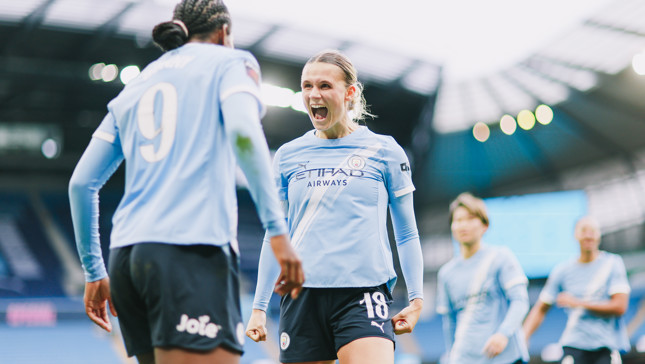 Women's football players celebrating on the field in Manchester City kits after scoring a goal at the Etihad Stadium. Visible numbers are 9 and 18. The stadium's roof is in the background.