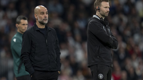 Two football coaches standing on the sideline during a match, with one wearing a headset. The crowd is blurred in the background.