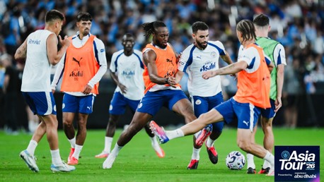 Manchester City players training during the Asia Tour 2026. The team members are wearing blue and white kits with orange training bibs. A logo indicates sponsorship by Revolut and Asahi. The event is titled Asia Tour '26 presented by Abu Dhabi.