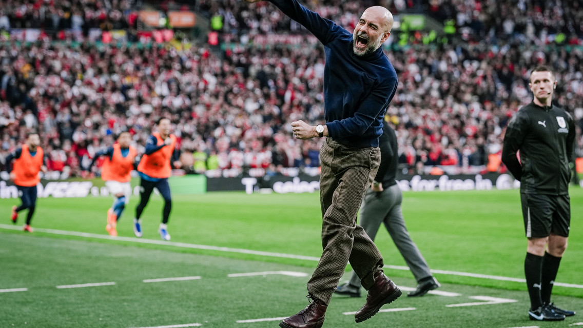 A soccer coach jumps in celebration on the sideline during a match, with players in orange bibs running in the background and a crowded stadium.