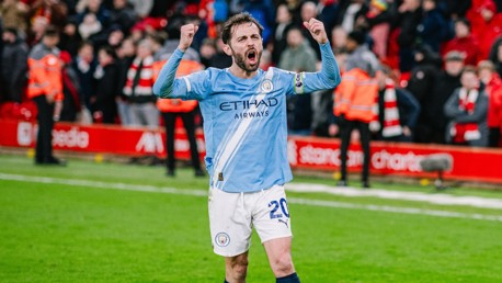 Soccer player in Manchester City kit celebrating on a football field with a crowd in the background.