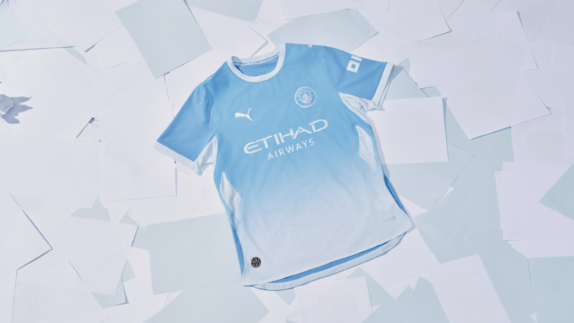 A Manchester City 2023-2024 home kit featuring light blue with sponsor Etihad Airways and Puma logos, laid on white paper sheets.