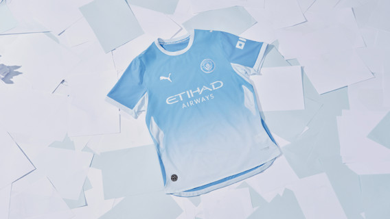 A Manchester City 2023-2024 home kit featuring light blue with sponsor Etihad Airways and Puma logos, laid on white paper sheets.