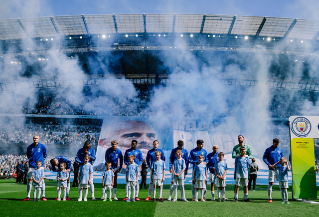 Manchester City team line up at Etihad Stadium for an event with smoke effects, banner, and players accompanied by young mascots.