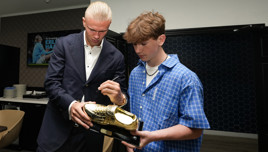 Two people are looking at a golden boot trophy, with one of them signing it. They're in a room with a television displaying the text 'An Evening with Erling Haaland'.