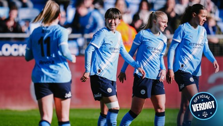 Manchester City women's soccer team players in light blue jerseys with Etihad Airways branding, celebrating on the field after a goal. The background shows fans in a stadium setting.