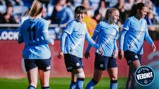 Manchester City women's soccer team players in light blue jerseys with Etihad Airways branding, celebrating on the field after a goal. The background shows fans in a stadium setting.