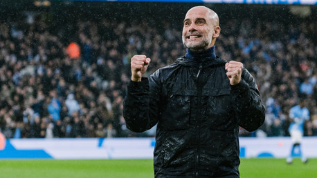 A man wearing a black jacket with his fists raised in celebration at a football stadium, surrounded by a crowd with blurred individuals in the background.