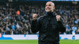 A man wearing a black jacket with his fists raised in celebration at a football stadium, surrounded by a crowd with blurred individuals in the background.