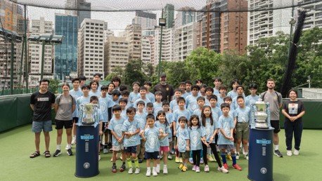 Group photo of children wearing light blue Manchester City shirts on a rooftop sports field, surrounded by coaches and trophies under netting with city buildings in the background.