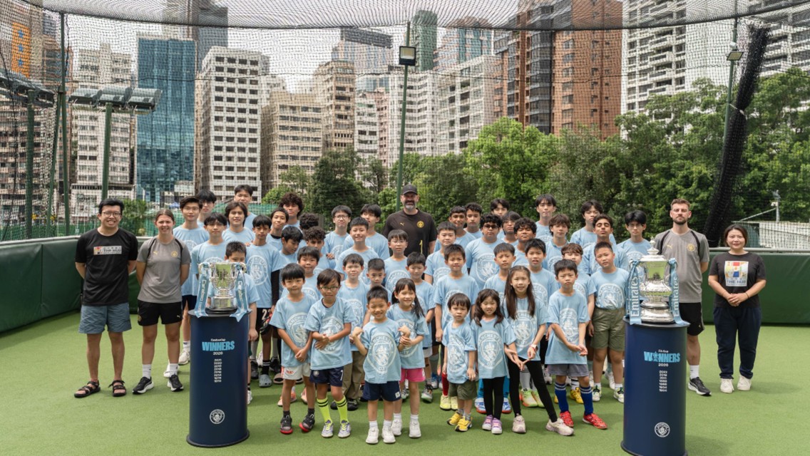 Group photo of children wearing light blue Manchester City shirts on a rooftop sports field, surrounded by coaches and trophies under netting with city buildings in the background.