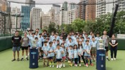Group photo of children wearing light blue Manchester City shirts on a rooftop sports field, surrounded by coaches and trophies under netting with city buildings in the background.