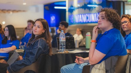 Individuals attending a conference or meeting in a room with 'The Mancunian' sign in the background.