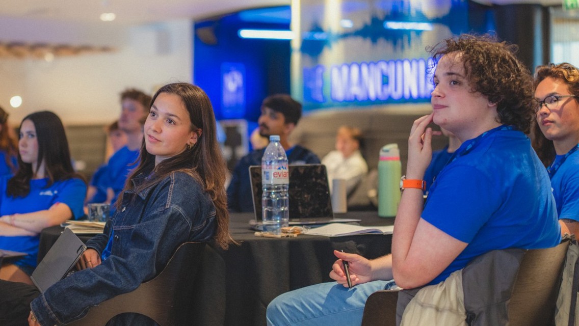 Individuals attending a conference or meeting in a room with 'The Mancunian' sign in the background.