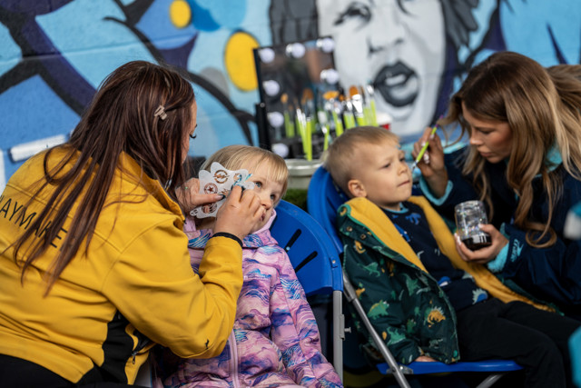 Children getting their faces painted at an event with colorful background and people collaborating.