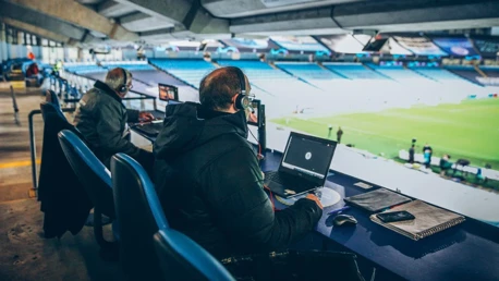 Two commentators with headphones and laptops in a stadium booth overlooking a field.