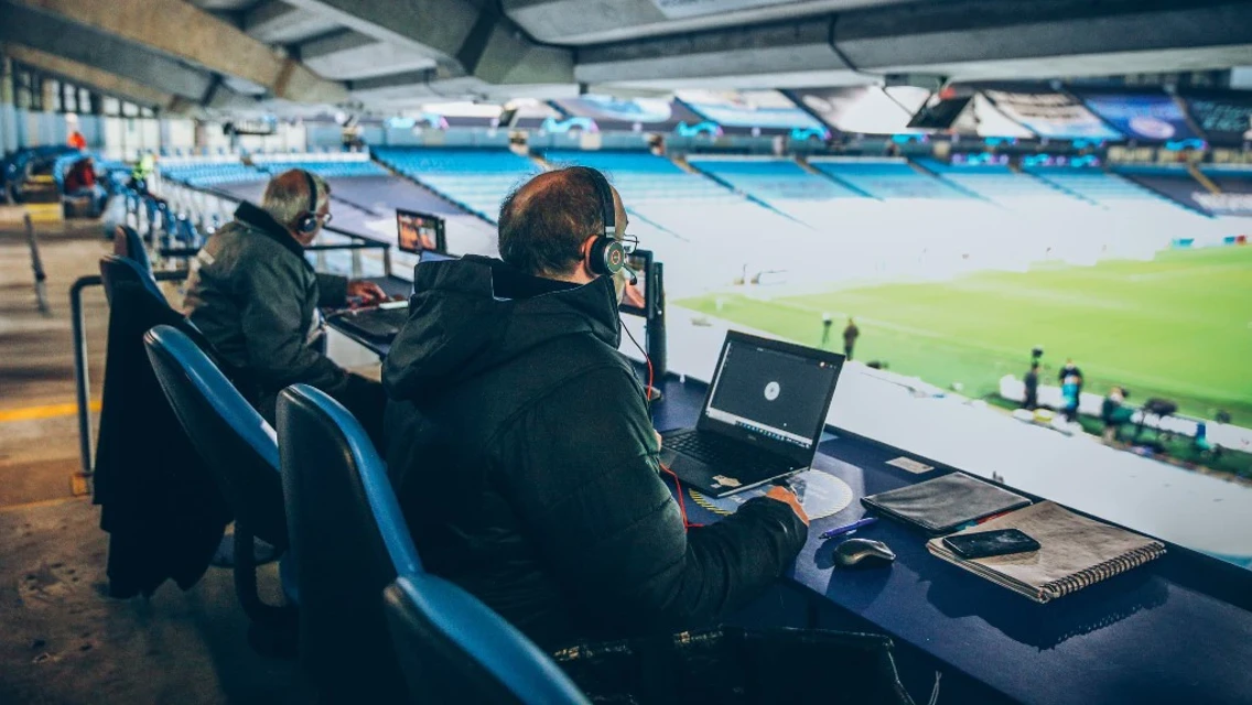 Two commentators with headphones and laptops in a stadium booth overlooking a field.