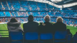Four people are seated in the stands of a football stadium, watching a game in progress. The seats in the background are primarily blue.