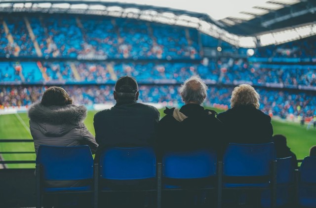 Four people are seated in the stands of a football stadium, watching a game in progress. The seats in the background are primarily blue.
