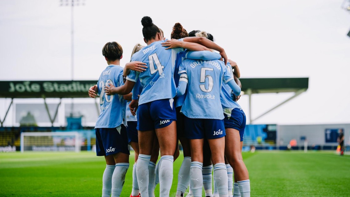 A group of soccer players in light blue jerseys huddling on the field at Joie Stadium with 'Revolut' and other text on their shirts.