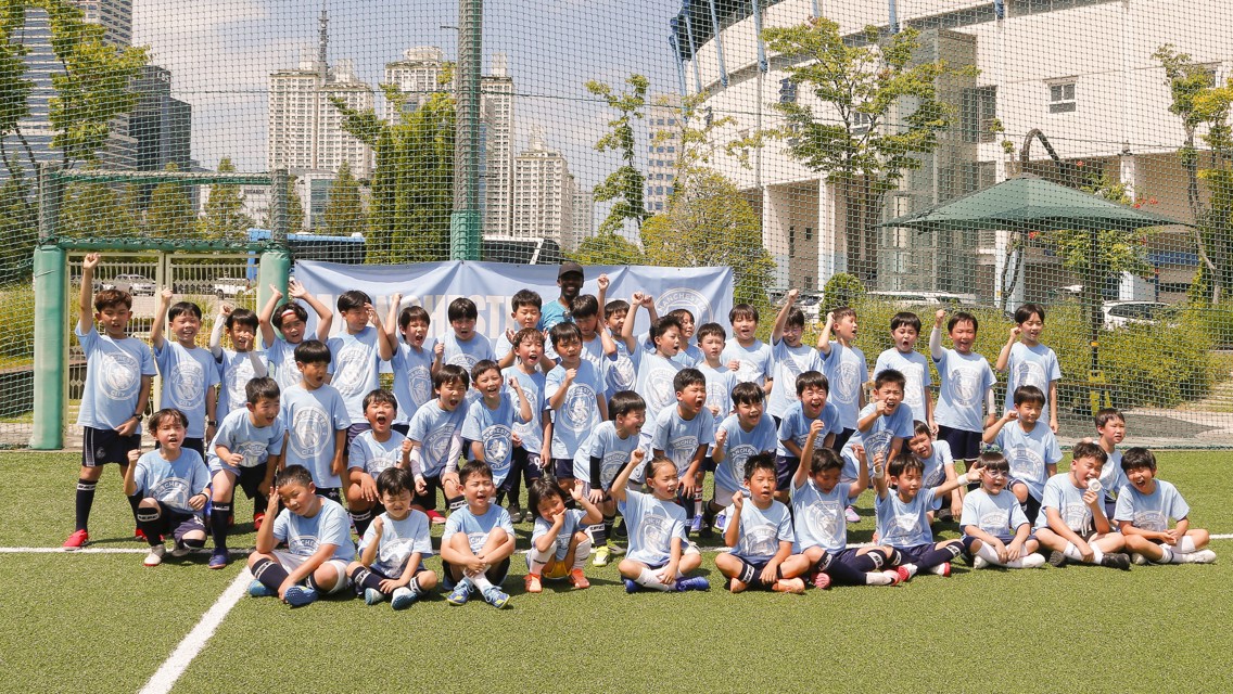 A group of young children are posing on a soccer field, all wearing matching light blue shirts with soccer-related logos. A banner is in the background, and buildings are visible beyond the fenced area.