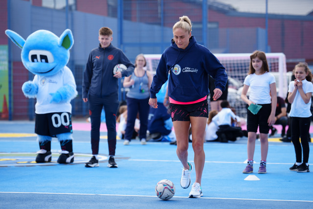 A Manchester City football coach is training children while the team's mascot and other participants are present on a sports court.
