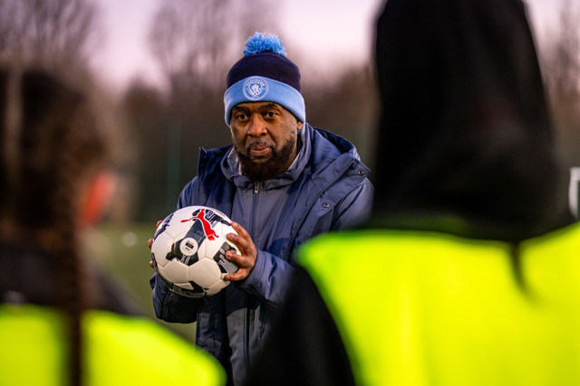 A football coach wearing a Manchester City hat is holding a football while addressing the team. The focus is on the coach holding the ball.