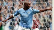 A football player in a Manchester City jersey celebrating a goal on the field. The jersey displays the Thomas Cook brand and the Man City crest.