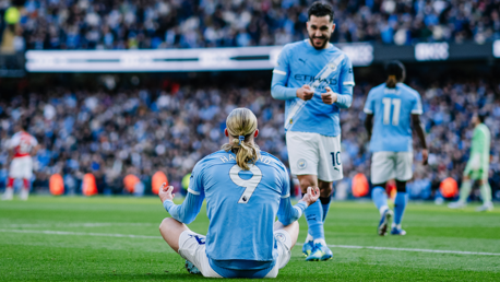 A Manchester City player sitting on the ground in a meditation pose after scoring, with teammates in blue kits around him and a crowd in the background.