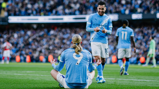A Manchester City player sitting on the ground in a meditation pose after scoring, with teammates in blue kits around him and a crowd in the background.