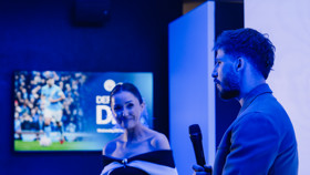 Two people on stage conversing with microphones; a football-related image on the screen in the background shows a player in a blue kit.