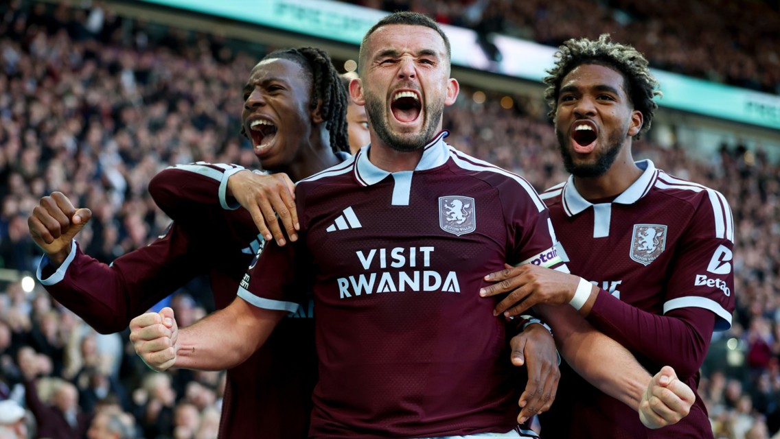 Three Aston Villa players celebrating a goal wearing maroon jerseys with 'Visit Rwanda' sponsor visible, in front of a cheering crowd.