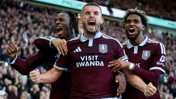 Three Aston Villa players celebrating a goal wearing maroon jerseys with 'Visit Rwanda' sponsor visible, in front of a cheering crowd.