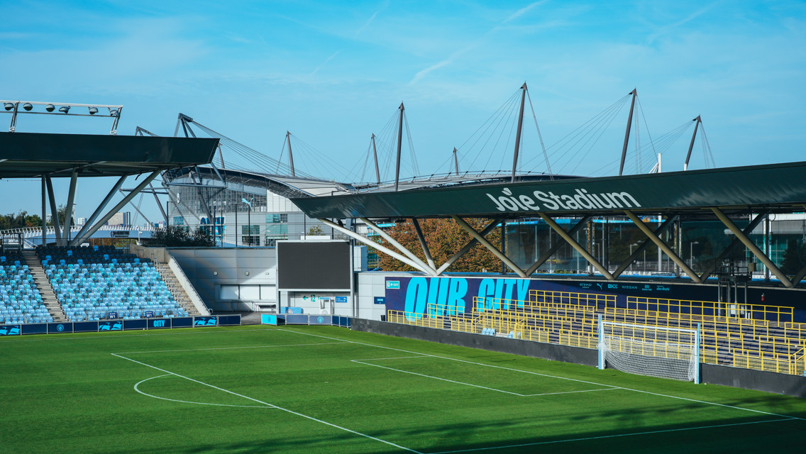 View of Joie Stadium with seating and goalposts in a football field, featuring the Etihad Stadium in the background.