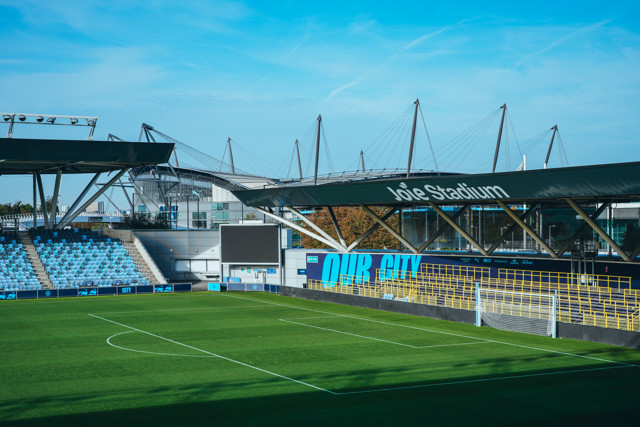 View of Joie Stadium with seating and goalposts in a football field, featuring the Etihad Stadium in the background.
