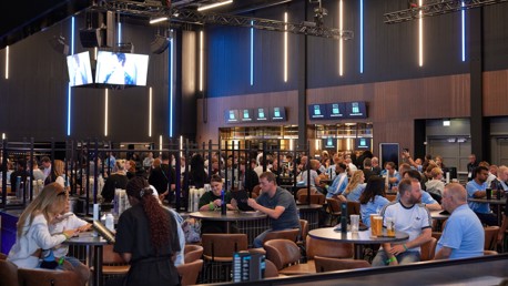 A bustling lounge area with people seated at tables in the Etihad Stadium. Screens displaying content and modern lighting create an energetic atmosphere.