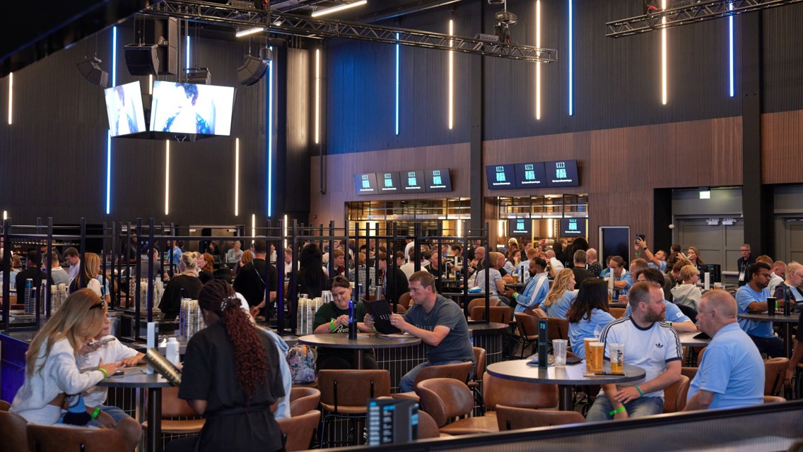 A bustling lounge area with people seated at tables in the Etihad Stadium. Screens displaying content and modern lighting create an energetic atmosphere.