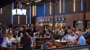 A bustling lounge area with people seated at tables in the Etihad Stadium. Screens displaying content and modern lighting create an energetic atmosphere.