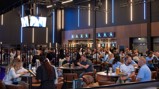 A bustling lounge area with people seated at tables in the Etihad Stadium. Screens displaying content and modern lighting create an energetic atmosphere.