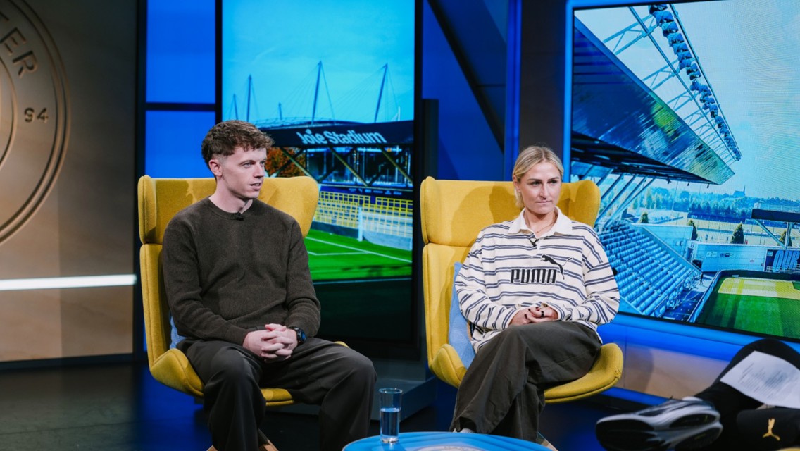 A man and a woman sitting in a studio. Yellow chairs, display showing 'Joie Stadium'.