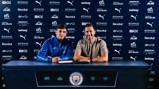 Two individuals sitting at a table with a Manchester City backdrop, signing a document. One wears a blue Manchester City tracksuit, the other casual attire.