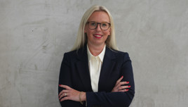 A woman with long blonde hair wearing a navy blue suit and white shirt, with arms crossed in front of a gray textured wall.