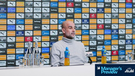 A person is seated at a press conference with a Manchester City sponsor backdrop. Bottles of water and a Lucozade drink are on the table.
